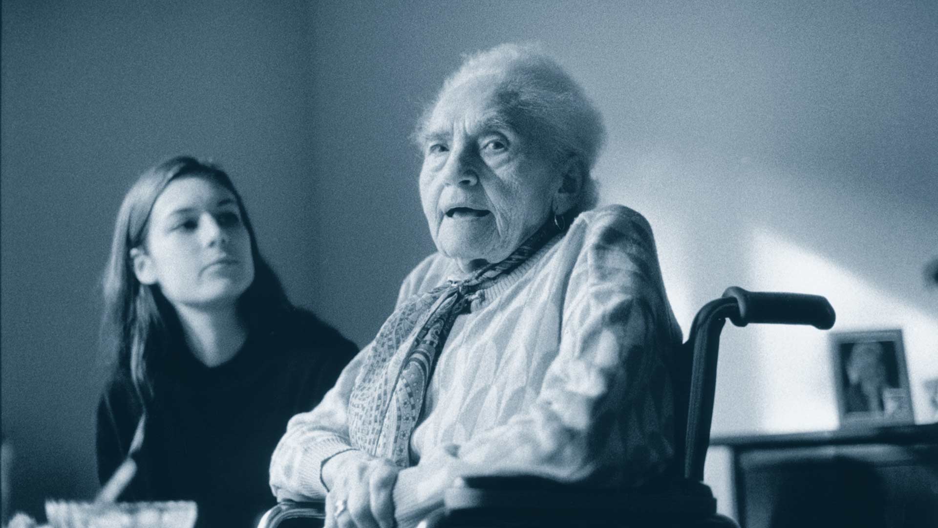 Im Vordergrund schaut eine ältere Frau in die Kamera, der Mund ist leicht geöffnet. Im Hintergrund schaut eine junge Frau die ältere Dame an.