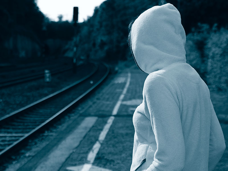 Eine Person mit Kapuzenpullover steht an einem Bahnhof und schaut entlang der Gleise